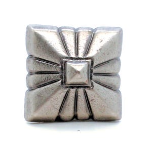 May include: A square silver-toned metal button featuring a raised central pyramid shape surrounded by radiating embossed lines that extend toward the four corners and edges, creating a geometric sunburst pattern with a metallic finish.