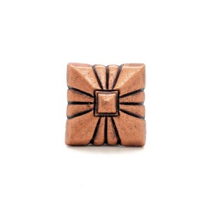 May include: A square copper-toned metal button featuring a raised central square surrounded by radiating embossed lines that extend toward the edges, creating a geometric sunburst pattern with a weathered, textured finish on the surface.