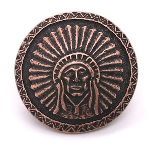 May include: A copper-colored metal button with a raised design of a Native American chief wearing a headdress. The button has a textured, circular border.