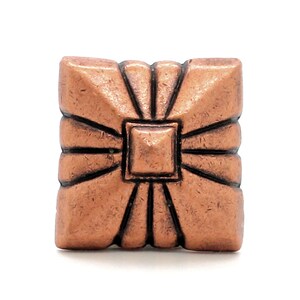 May include: A square-shaped button with a metallic copper finish. The surface features a raised central square surrounded by radiating lines that extend toward the edges, creating a textured, geometric pattern with dark recessed grooves.