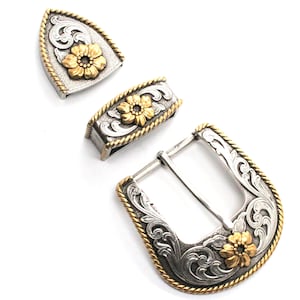 May include: A silver and gold-tone western belt buckle set. The set includes a buckle, a belt loop, and a tip. Each piece features a gold flower and scroll design. The edges are trimmed with a gold rope design.