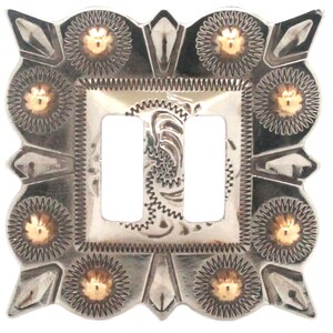 May include: A silver-toned, decorative light switch cover with two rectangular openings. The cover features intricate embossed patterns, including floral and geometric designs, and small gold-colored accents. The overall design has a Western or Southwestern aesthetic.