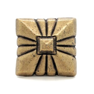 May include: A square-shaped metal button with an antique brass finish. The surface features a raised central square surrounded by radiating embossed lines that extend toward the edges, creating a sunburst pattern across the entire face.