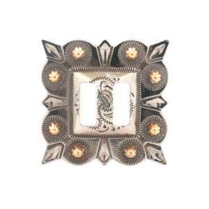 May include: Silver-toned metal light switch cover with two rectangular openings. The cover features a square central design with an ornate pattern and gold-colored accents. The edges are decorated with gold-colored sunburst designs.