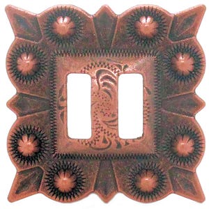 May include: A copper-colored, square light switch cover with two rectangular openings. The cover features an ornate, embossed design with floral and geometric patterns. The edges are scalloped with decorative elements.