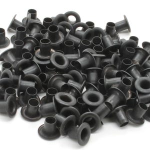 May include: A pile of black metal eyelets, each with a circular eye and a flared base. The eyelets are scattered, with varying orientations, against a white background. The eyelets are likely for crafting or industrial use.