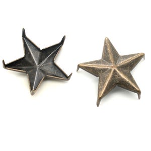May include: Two five-pointed star-shaped metal studs. One stud has a dark, gunmetal grey finish with prongs facing upward, while the other has a distressed bronze finish with prongs facing downward, showing the textured surface of the metal.
