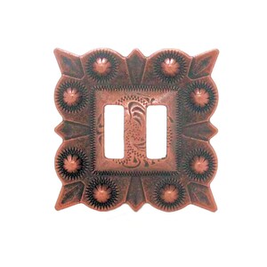 May include: A copper-colored, square belt buckle with an ornate, floral design. The buckle has two rectangular openings in the center and is surrounded by a detailed, scalloped edge with sunburst-like accents. The overall design has a Western or Southwestern aesthetic.