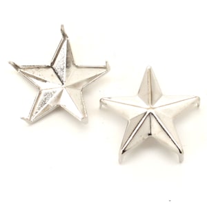 May include: Two silver-colored metal star-shaped studs with five points. Each star features a raised center and small prongs extending from the tips of the points, designed for attachment to fabric or other materials. The surface is smooth and reflective.
