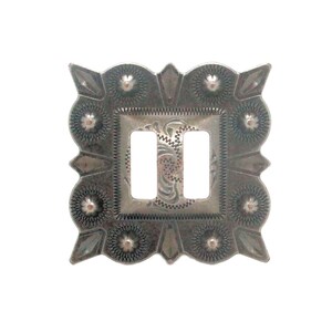 May include: A silver-toned metal buckle with a square central design featuring two rectangular openings. The buckle has an ornate, textured border with floral and geometric patterns. The overall design is symmetrical and detailed.