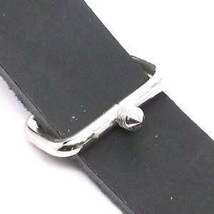 Puede incluir: Primer plano de una hebilla de correa de reloj plateada con una punta puntiaguda. La hebilla est&aacute; unida a una correa de reloj de cuero negro.