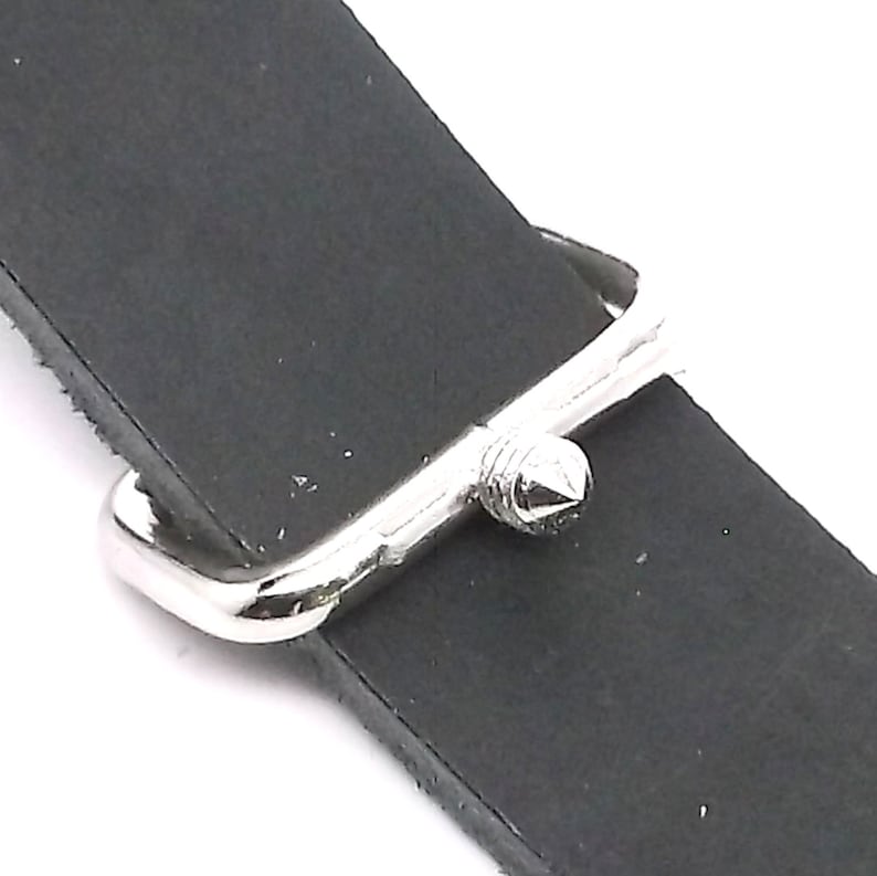 Puede incluir: Primer plano de una hebilla de correa de reloj plateada con una punta puntiaguda. La hebilla est&aacute; unida a una correa de reloj de cuero negro.