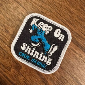 A rectangular embroidered patch features the phrase "Keep On Shining" in white letters above a blue cartoon character dancing. The background is black with a white border, and the lower section reads "Okie Shine" in blue letters.