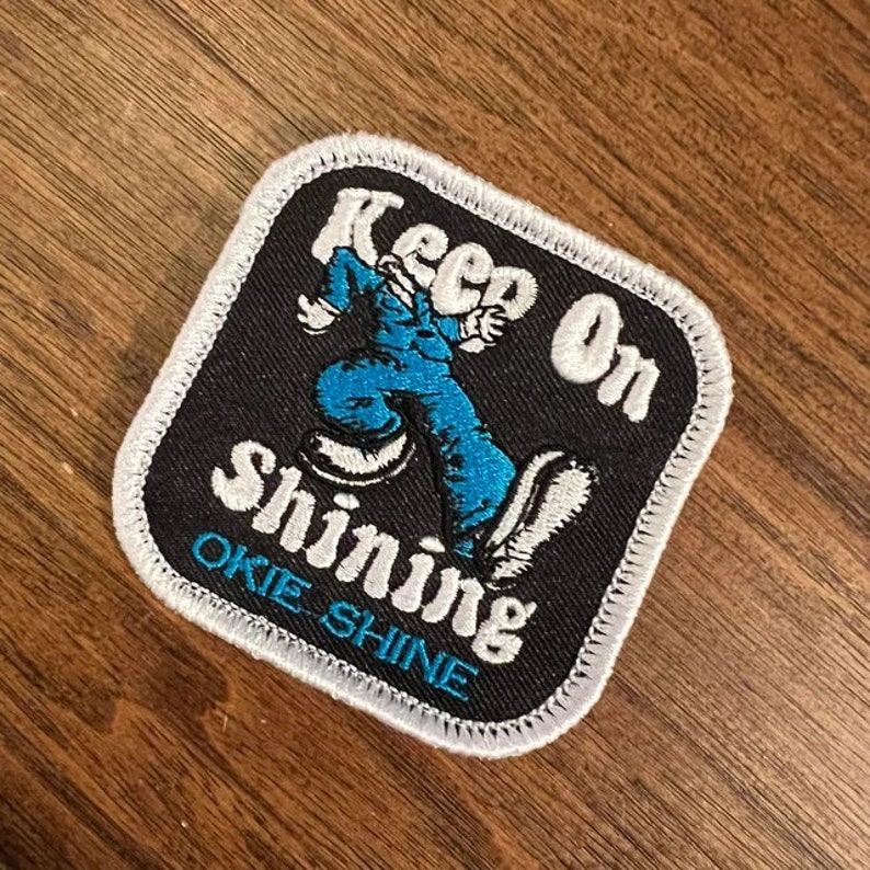 A rectangular embroidered patch features the phrase "Keep On Shining" in white letters above a blue cartoon character dancing. The background is black with a white border, and the lower section reads "Okie Shine" in blue letters.