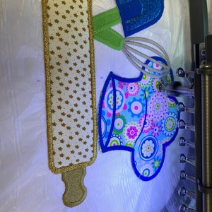 A vibrant embroidered kitchen scene featuring a colorful apron with floral patterns, a golden rolling pin, and a blue whisk. The backdrop is illuminated, showcasing the intricate details of the embroidery.