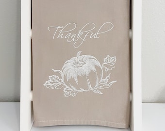 May include: A beige kitchen towel featuring the word "Thankful" in cursive and a pumpkin design. The towel is displayed on a white wooden ladder rack, ideal for autumn. The towel is approximately 40 cm in length.