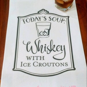 May include: White kitchen towel with black text that reads "Today's Soup: Whiskey with Ice Croutons". A glass of whiskey with ice is pictured above the text.