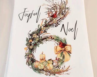 May include: A white rectangular kitchen towel featuring a printed illustration of a wreath made of twigs, pine needles, and berries. The wreath includes a lit candle, ornaments, a large tan bow, and two red cardinals. The text Joyful Noel is printed in black.
