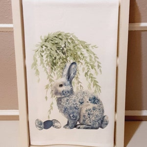May include: A white rectangular kitchen towel featuring a printed illustration of a rabbit with a blue and white floral pattern on its body. The rabbit is positioned under a green willow tree with three small decorative eggs placed nearby.