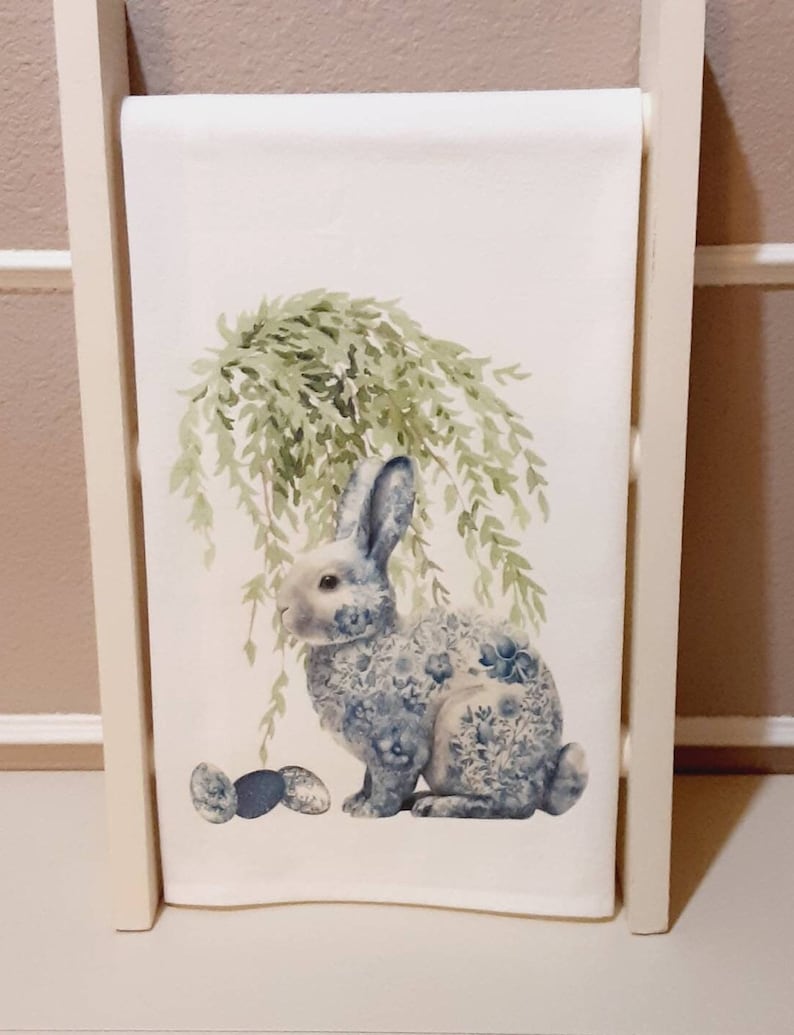 May include: A white rectangular kitchen towel featuring a printed illustration of a rabbit with a blue and white floral pattern on its body. The rabbit is positioned under a green willow tree with three small decorative eggs placed nearby.