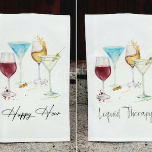 May include: Two white rectangular kitchen towels featuring watercolor illustrations of four cocktail glasses filled with various drinks and olives. The left towel displays the text Happy Hour, while the right towel displays the text Liquid Therapy.