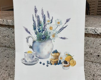 May include: A white tea towel featuring a watercolour illustration of a jug filled with lavender flowers, daisies, and blueberries. The jug is placed on a table with a teacup, lemon slices, and blueberry muffins. The text "Idle Tyme Creations" is printed in the bottom right corner.