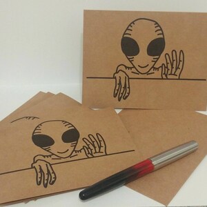 May include: A stack of rectangular brown kraft paper cards featuring a black line drawing of an alien peeking over a horizontal line. The alien has large oval eyes and a thin hand raised in a wave. A fountain pen with a red and black barrel is present.
