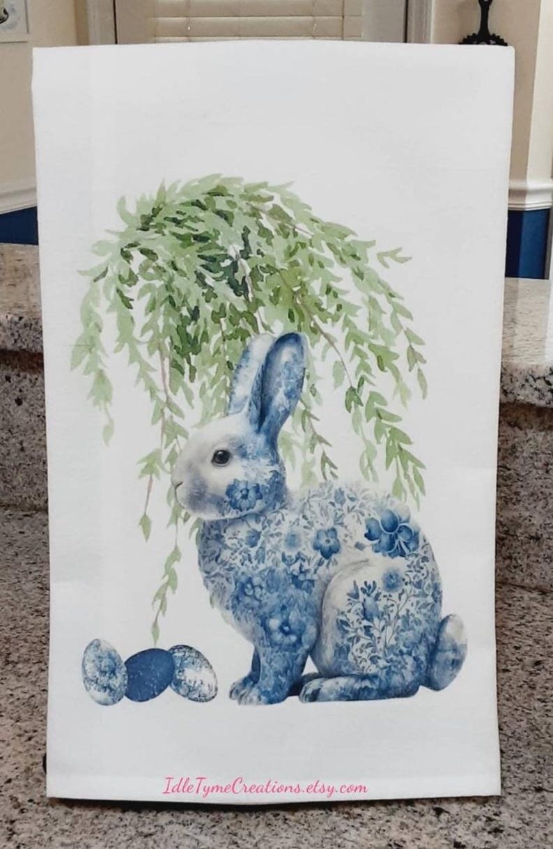 May include: A white rectangular kitchen towel featuring a watercolour illustration of a rabbit with a blue and white floral pattern. Above the rabbit hangs green willow foliage, and three blue speckled eggs sit at its feet. IdleTymeCreations.etsy.com