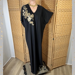 May include: Black maxi dress with gold floral embroidery on the bodice. The dress features a V-neck and short sleeves. A gold-tone necklace is worn on a mannequin. The dress is displayed in front of a woven room divider.