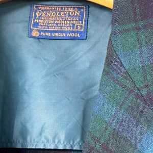 Puede incluir: Primer plano de una chaqueta Pendleton con un estampado de cuadros azules y verdes. El forro interior es turquesa. Una etiqueta azul dice "WARRANTED TO BE A PENDLETON" y "PURE VIRGIN WOOL". La chaqueta es talla S.