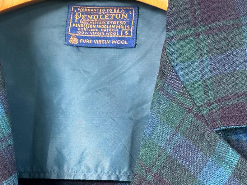 Puede incluir: Primer plano de una chaqueta Pendleton con un estampado de cuadros azules y verdes. El forro interior es turquesa. Una etiqueta azul dice "WARRANTED TO BE A PENDLETON" y "PURE VIRGIN WOOL". La chaqueta es talla S.
