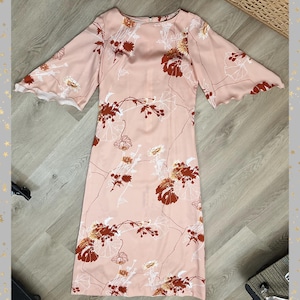 May include: A light pink dress with a floral pattern featuring brown and white flowers and stems. The dress has wide, elbow-length sleeves with a scalloped edge. The dress is knee-length.
