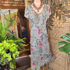May include: A gray dress with a floral pattern featuring pink roses and green leaves. The dress has short sleeves and a V-neckline. A decorative necklace is around the mannequin's neck. The dress is displayed in front of a woven background.