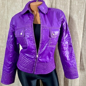 May include: A purple shiny jacket with a zipper closure and two flap pockets. The jacket has a quilted design and a stand-up collar.