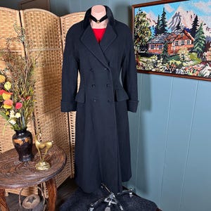 May include: A long, black, double-breasted coat featuring a wide collar, long sleeves with cuffs, and a fitted waistline. The coat is displayed on a mannequin, showing a red garment underneath and a black choker necklace around the neck area.