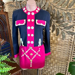 May include: A two-piece set consisting of a navy blue long-sleeve jacket and a matching hot pink skirt. The jacket features a hot pink center panel, two flap pockets, and round white buttons. The skirt is adorned with a geometric button pattern.