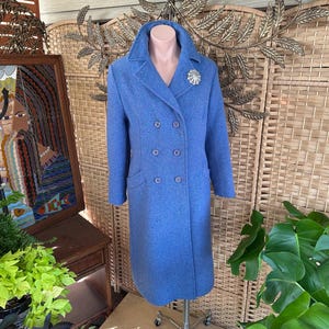 May include: A long, double-breasted, blue coat with a notched collar and a decorative brooch. The coat has two rows of buttons and two front pockets. The coat is displayed on a mannequin.
