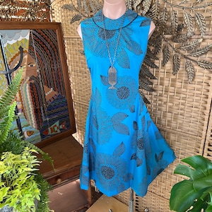 May include: A sleeveless, turquoise dress with a black floral pattern. The dress has a round collar and a flared skirt. A silver-toned pendant hangs from a necklace. The dress is displayed on a mannequin.
