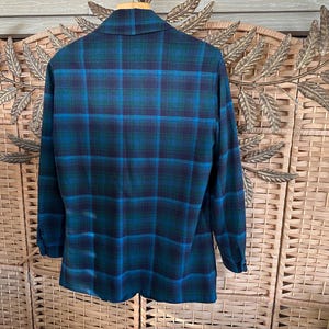 Puede incluir: Blazer a cuadros de manga larga en tonos turquesa, azul y marino. La parte trasera de la chaqueta es visible, mostrando el cl&aacute;sico estampado tart&aacute;n. El blazer tiene cuello y pu&ntilde;os abotonados.