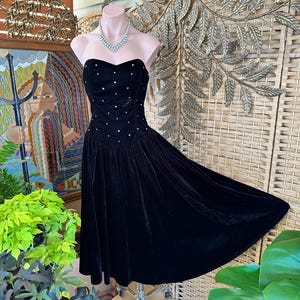 May include: A strapless black velvet dress with a sweetheart neckline and a full skirt. The bodice is embellished with small, scattered white rhinestones. The dress is displayed on a mannequin, showcasing its elegant design and vintage style.