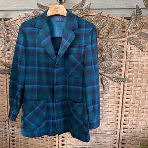 Puede incluir: Un blazer vintage a cuadros en tonos turquesa, azul y negro. La chaqueta presenta un cuello con solapas cl&aacute;sico, tres botones y dos bolsillos delanteros. El blazer est&aacute; colgado de una percha de madera.