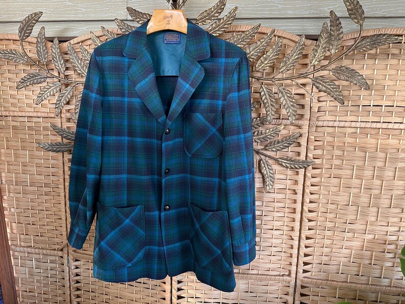 Puede incluir: Un blazer vintage a cuadros en tonos turquesa, azul y negro. La chaqueta presenta un cuello con solapas cl&aacute;sico, tres botones y dos bolsillos delanteros. El blazer est&aacute; colgado de una percha de madera.