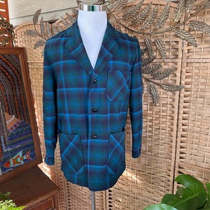 Puede incluir: Un blazer vintage a cuadros en tonos azul y verde, exhibido en un maniqu&iacute;. La chaqueta presenta un cuello con solapas, tres botones y dos bolsillos delanteros. El patr&oacute;n de cuadros es un dise&ntilde;o llamativo.