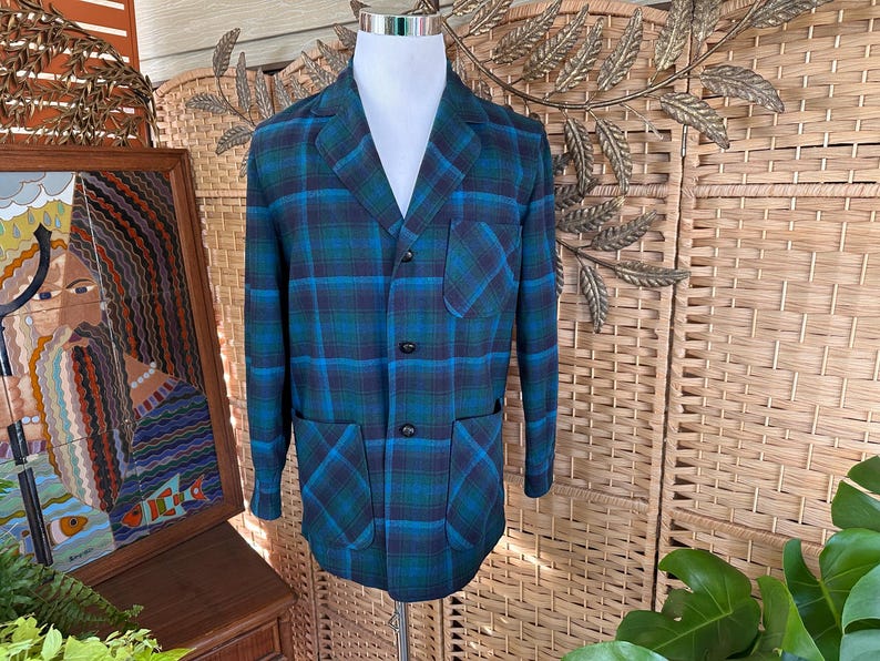 Puede incluir: Un blazer vintage a cuadros en tonos azul y verde, exhibido en un maniqu&iacute;. La chaqueta presenta un cuello con solapas, tres botones y dos bolsillos delanteros. El patr&oacute;n de cuadros es un dise&ntilde;o llamativo.