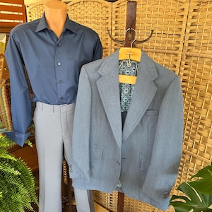 May include: A blue button-down shirt and gray checkered pants are displayed on a mannequin. A light blue blazer hangs on a wooden hanger. The blazer has two front pockets and a patterned lining. The outfit is displayed in front of a woven background.