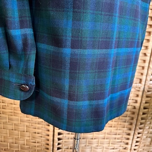 Puede incluir: Primer plano de una tela de cuadros verde azulado, azul y marino. La tela es un material tejido con un patr&oacute;n de cuadros cl&aacute;sico. El pu&ntilde;o de la manga con un bot&oacute;n marr&oacute;n es visible en el lado izquierdo. La tela est&aacute; drapeada sobre un soporte.