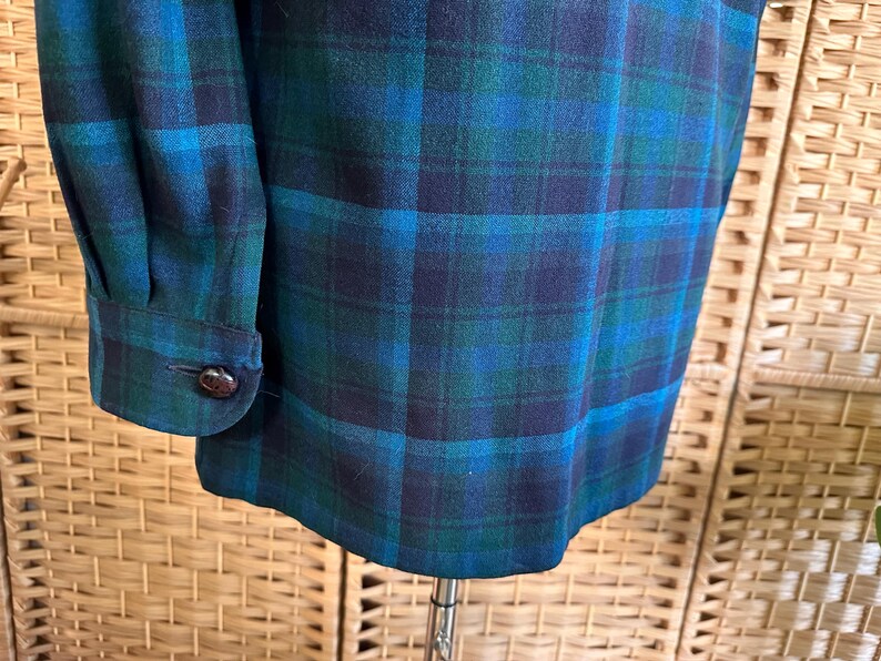 Puede incluir: Primer plano de una tela de cuadros verde azulado, azul y marino. La tela es un material tejido con un patr&oacute;n de cuadros cl&aacute;sico. El pu&ntilde;o de la manga con un bot&oacute;n marr&oacute;n es visible en el lado izquierdo. La tela est&aacute; drapeada sobre un soporte.