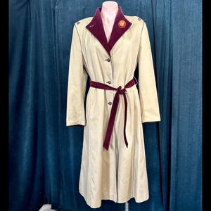 May include: A beige trench coat with a burgundy velvet collar and belt. The coat features a button-down front, long sleeves, and a decorative pin on the collar. The coat is displayed on a mannequin.