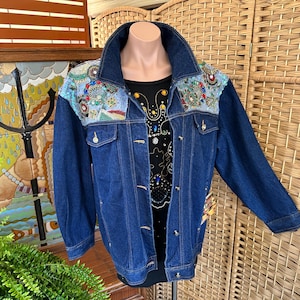 May include: A blue denim jacket with decorative embroidery on the shoulders. The jacket has a classic collar, button closures, and pockets. A black shirt with a colorful design is visible underneath.