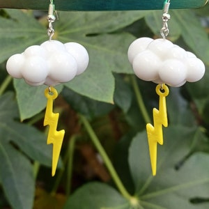 May include: A pair of earrings featuring white cloud charms with yellow lightning bolt accents. The earrings are hanging from silver hooks.